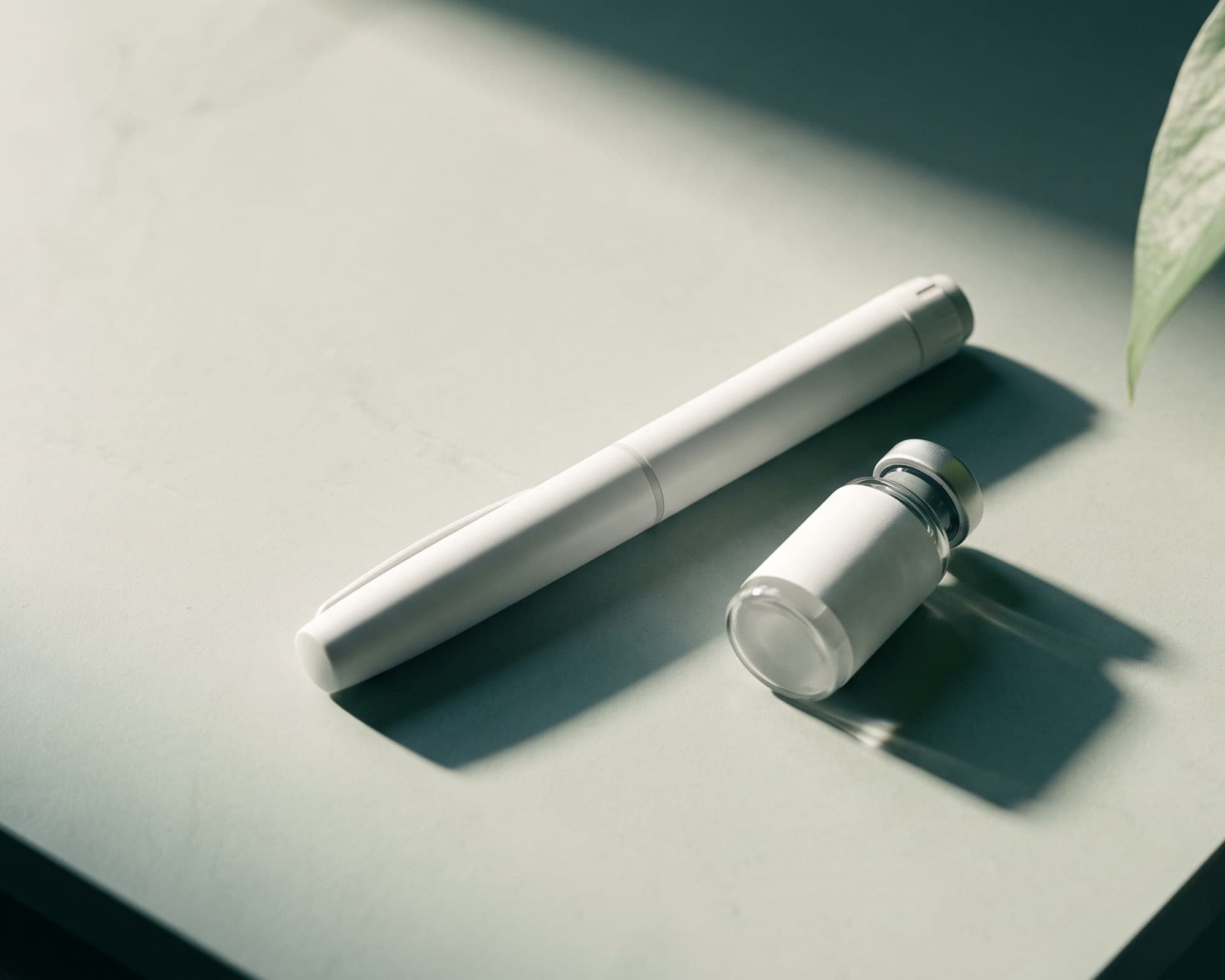 An unlabelled injection pen and a small vial on a pale stone surface beside a plant leaf.