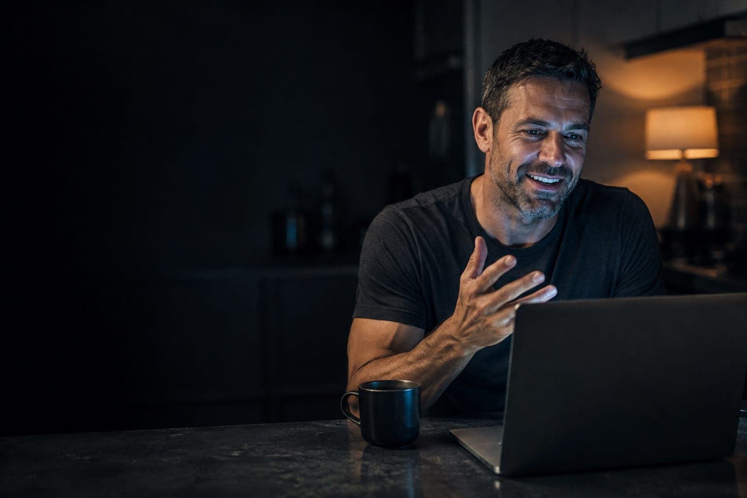 A man in his forties leaning into a video call at his kitchen counter in the evening, talking and smiling.