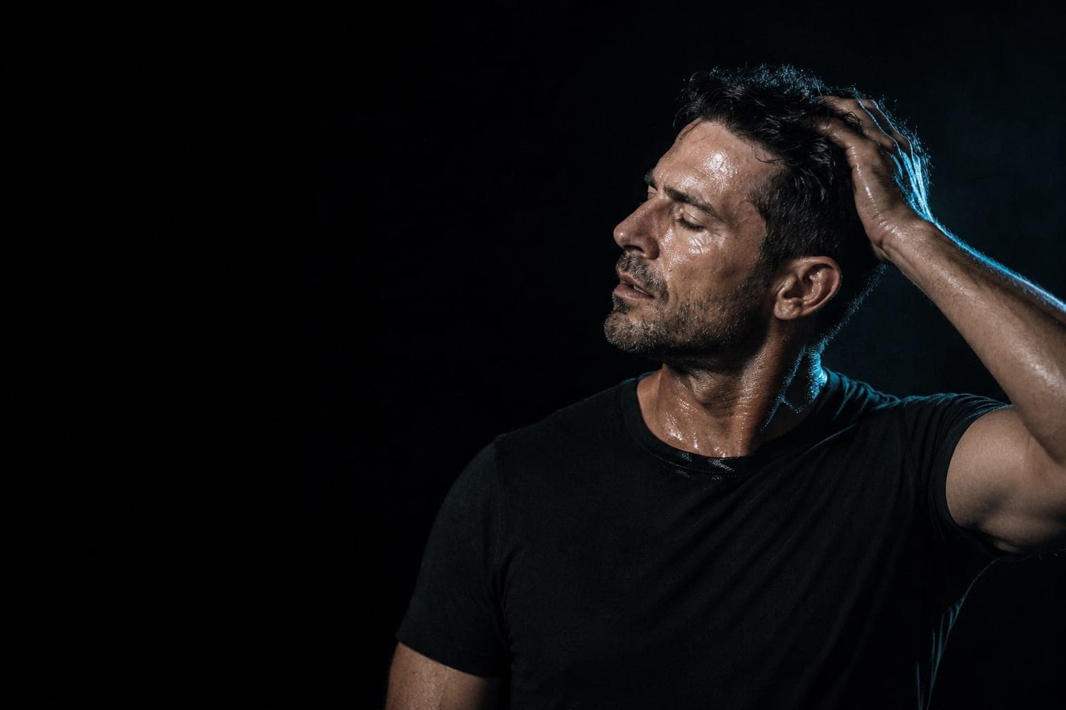 A man in his forties running a hand back through damp hair, lit hard from one side against a dark background.