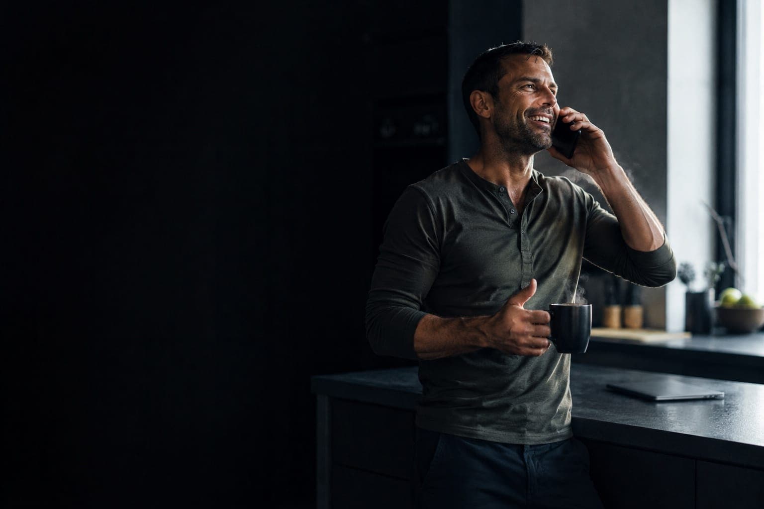 A man in his forties laughing on a phone call at his kitchen counter, a mug in his other hand.