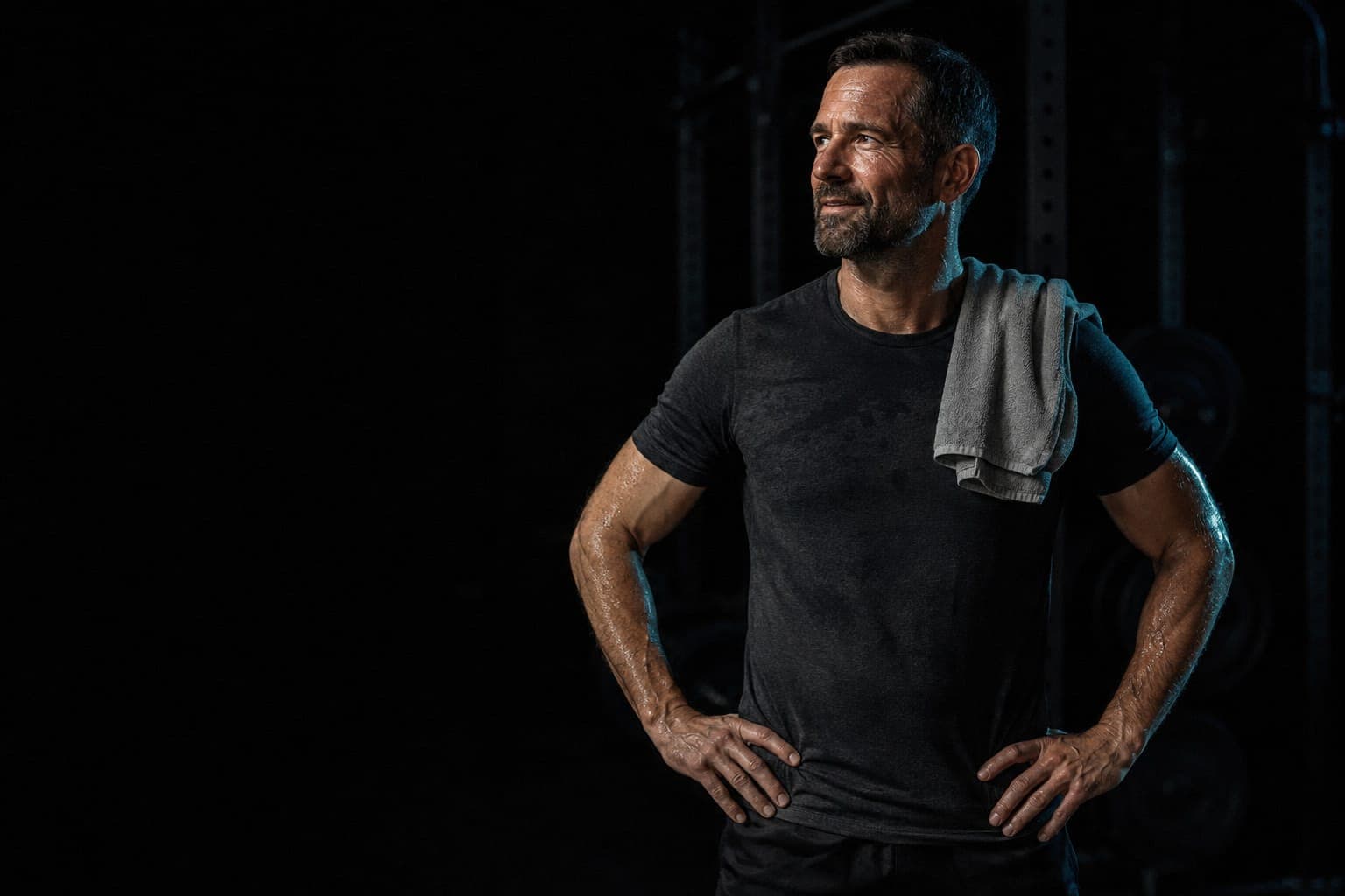 A man in his forties standing in a dark home gym after training, a towel over one shoulder, hands on his hips.